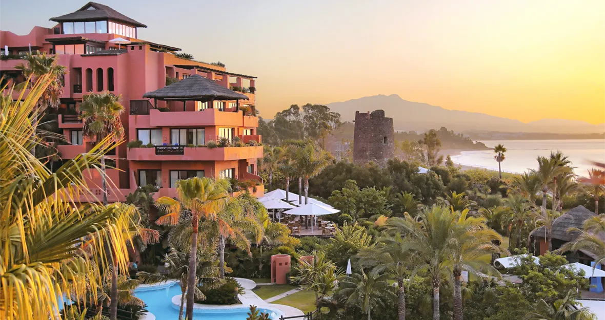 Das Melia Hacienda del Conde, ein Resort in leuchtendem Rosa mit üppigen Palmen und einem gewundenen Pool, bietet bei Sonnenuntergang einen Blick auf die malerische Küste, wobei im Hintergrund die Berge und das Meer zu sehen sind.