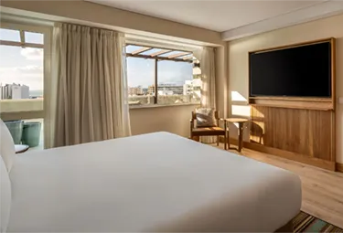 Ein helles Hotelzimmer im MiM Sotogrande Meliá mit einem großen Bett, einem Holzstuhl und einem kleinen Tisch am Fenster mit Blick auf die Stadt, hellen Vorhängen und einem an der Wand montierten Flachbildfernseher.