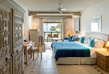 Ein Hotelzimmer mit zwei Betten, blauer Bettwäsche, Holzmöbeln, einem Schreibtisch, Lampen und einem Balkon mit Blick auf das Meer und die Palmen – ideal zum Entspannen nach Ihrer Golfrundreise durch Irland. Das Zimmer ist hell und ordentlich eingerichtet.