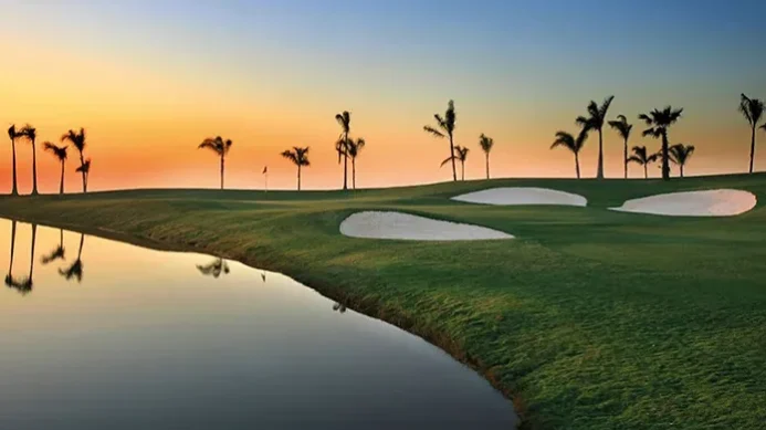 Ein Golfplatz bei Sonnenuntergang mit Palmen, Sandbunkern und einer Flagge in der Ferne. Ein ruhiges Wasserhindernis spiegelt den farbenprächtigen Himmel wider – eine Szene, die an Momente auf einer Golfrundreise durch Irland erinnert.