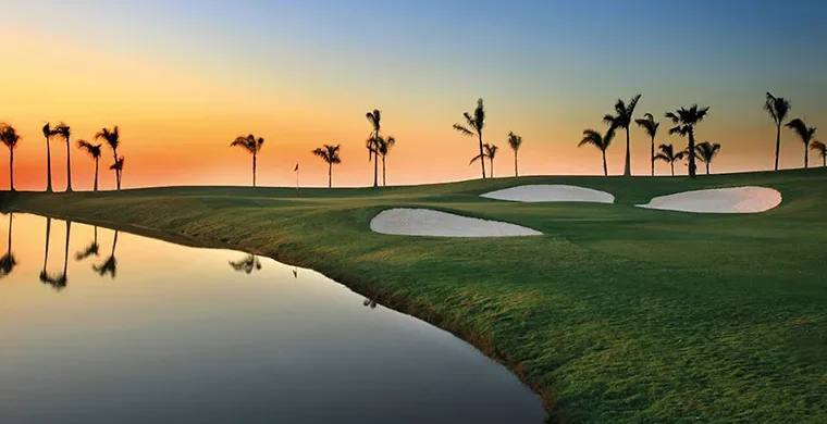 Kairo-Allegria-GC Ein Golfplatz bei Sonnenuntergang mit Palmen, Sandbunkern und einer Flagge in der Ferne. Ein ruhiges Wasserhindernis spiegelt den farbenprächtigen Himmel wider – eine Szene, die an Momente auf einer Golfrundreise durch Irland erinnert.