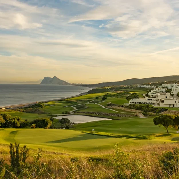 Ein malerischer Golfplatz an der Küste mit grünen Fairways und Sandbunkern, gesäumt von Bäumen, in der Nähe des eleganten Hotels „Melia Hacienda del Conde“ gelegen, mit weißen Gebäuden am Hang und einer felsigen Halbinsel in der Ferne unter einem teilweise bewölkten Abendhimmel.