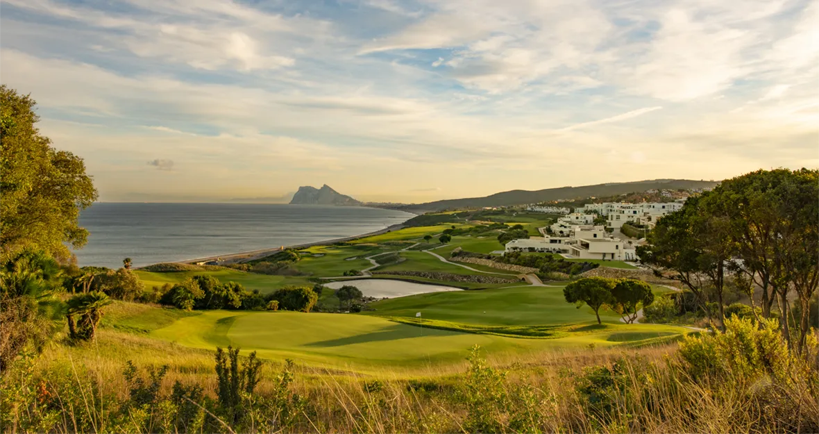 Ein malerischer Golfplatz an der Küste mit grünen Fairways und Sandbunkern, gesäumt von Bäumen, in der Nähe des eleganten Hotels „Melia Hacienda del Conde“ gelegen, mit weißen Gebäuden am Hang und einer felsigen Halbinsel in der Ferne unter einem teilweise bewölkten Abendhimmel.