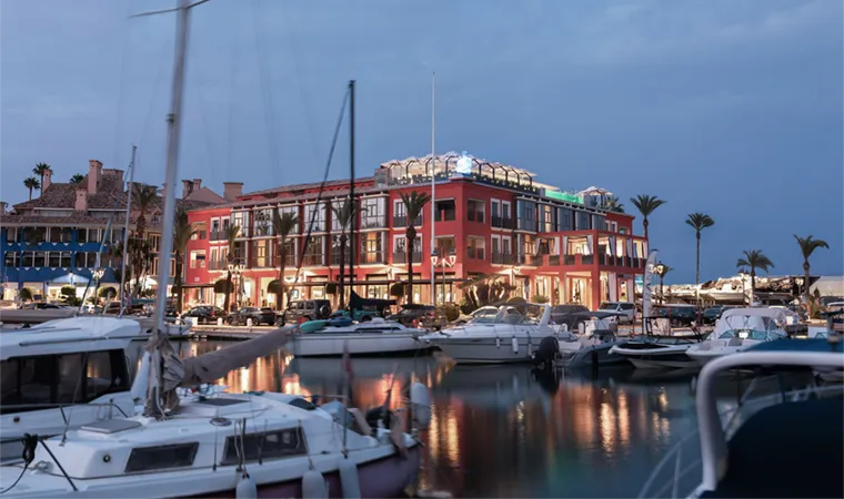 Ein Yachthafen in der Abenddämmerung mit mehreren vor Anker liegenden Booten im Vordergrund. Im Hintergrund sticht unter dem bewölkten Himmel das hell erleuchtete „Melia Hacienda del Conde“ hervor, ein rotes, dreistöckiges Gebäude mit großen Fenstern und Palmen in der Nähe.