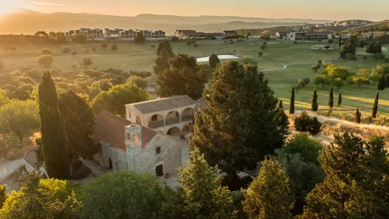Luftaufnahme eines von Bäumen umgebenen Steingebäudes inmitten einer grünen Landschaft mit Golfplatz – eine ideale Kulisse für eine Golfrundreise durch Irland – sowie moderne Gebäude in der Ferne bei Sonnenuntergang.