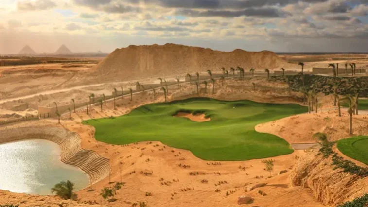 Inmitten einer sandigen Wüstenlandschaft sticht ein üppig grüner Golfplatz hervor, dessen Fairway von Palmen gesäumt ist. Im Hintergrund sind unter einem teilweise bewölkten Himmel die Pyramiden schemenhaft zu erkennen – ein exotischer Kontrast zu jeder Golfrundreise durch Irland.