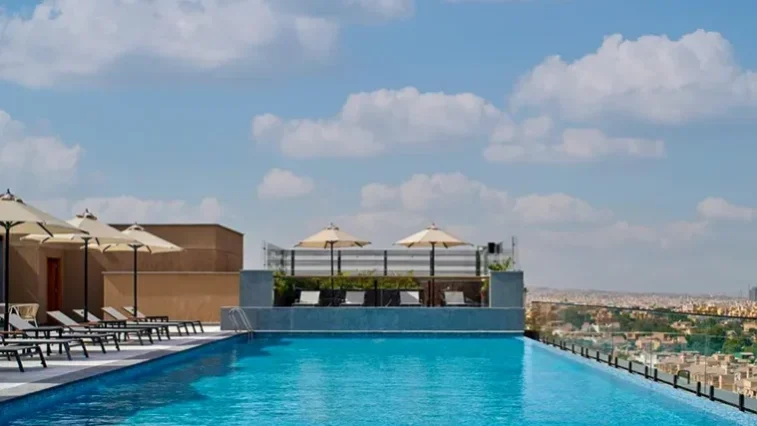 Ein Infinity-Pool auf dem Dach mit Liegestühlen und Sonnenschirmen, ideal zum Entspannen nach Ihrer Golfrundreise durch Irland, mit Blick auf die Stadtlandschaft unter einem teilweise bewölkten Himmel.