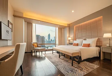 Ein modernes Hotelzimmer mit einem großen Bett, zwei Nachttischen, einem Schreibtisch mit Stuhl, einem Sessel am Fenster und einem Blick auf die Skyline der Stadt durch das raumhohe Fenster. Warme Beleuchtung sorgt für eine gemütliche Atmosphäre.
