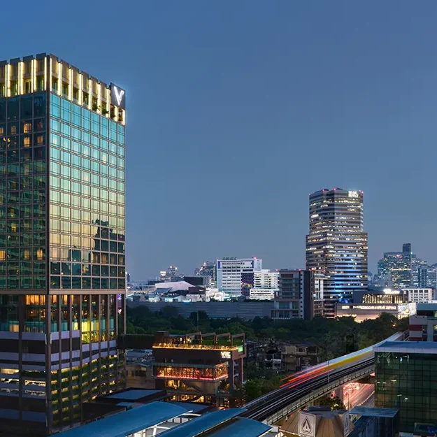 Eine Stadt-Skyline in der Abenddämmerung mit modernen Hochhäusern, darunter ein hohes Glasgebäude mit beleuchteten Fenstern auf der linken Seite und weitere beleuchtete Wolkenkratzer im Hintergrund vor einem blauen Abendhimmel.