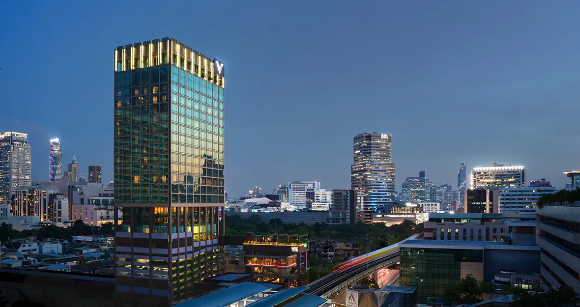 Eine Stadt-Skyline in der Abenddämmerung mit modernen Hochhäusern, darunter ein hohes Glasgebäude mit beleuchteten Fenstern auf der linken Seite und weitere beleuchtete Wolkenkratzer im Hintergrund vor einem blauen Abendhimmel.
