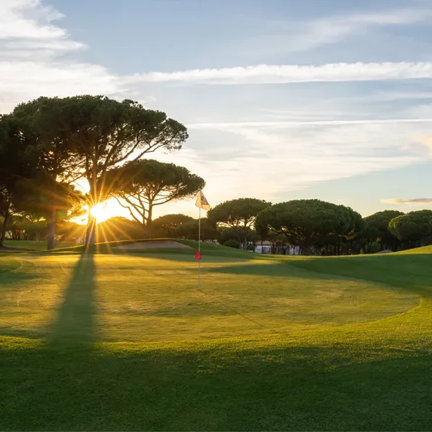 Golfplatz bei Sonnenuntergang, wobei das Sonnenlicht durch die Bäume scheint und lange Schatten auf das Grün des MiM Sotogrande Meliá wirft. Der Flaggenstock steht in der Nähe des Lochs, und rechts ist unter einem teilweise bewölkten Himmel ein Sandbunker zu sehen.
