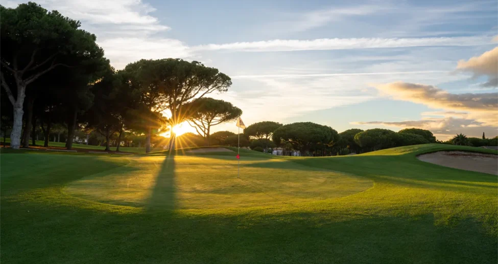 Golfplatz bei Sonnenuntergang, wobei das Sonnenlicht durch die Bäume scheint und lange Schatten auf das Grün des MiM Sotogrande Meliá wirft. Der Flaggenstock steht in der Nähe des Lochs, und rechts ist unter einem teilweise bewölkten Himmel ein Sandbunker zu sehen.