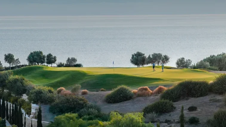 Ein Golfplatzgrün an einem Hang mit Blick auf das ruhige Meer, auf dem zwei Personen in der Nähe der Fahne stehen. Umgeben von Bäumen und Sträuchern unter einem klaren Himmel ist dies die perfekte Kulisse für ein unvergessliches Abenteuer auf einer Golfrundreise durch Irland.