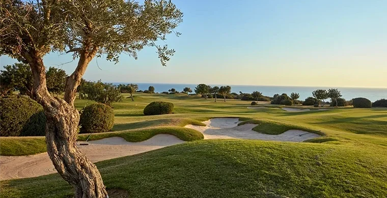 Ein malerischer Golfplatz mit Sandbunkern, saftig grünem Gras und Bäumen, mit Blick auf das Meer unter strahlend blauem Himmel bei Sonnenuntergang – perfekt für ein Abenteuer auf einer Golfrundreise durch Zypern.
