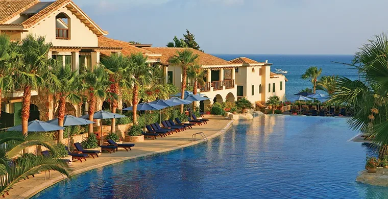 Ein luxuriöses Resort mit Palmen, Liegestühlen und blauen Sonnenschirmen, die einen großen Pool säumen, mit Blick auf das Meer und Boote in der Ferne unter strahlend blauem Himmel – ideal zum Entspannen nach Ihrer Golfrundreise durch Irland.