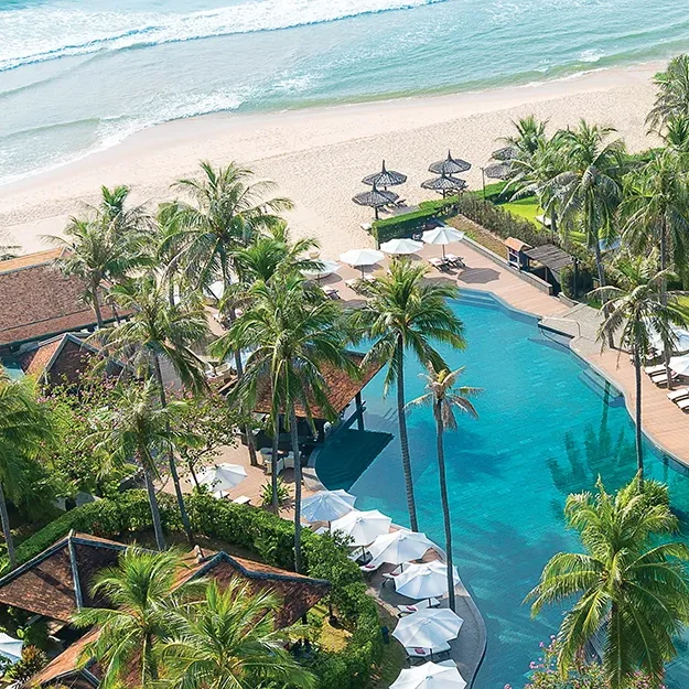 Luftaufnahme eines tropischen Strandresorts mit einem großen Pool, umgeben von Palmen, Liegestühlen, Sonnenschirmen und Gebäuden im traditionellen Stil, direkt an einem Sandstrand und den Wellen des Meeres.