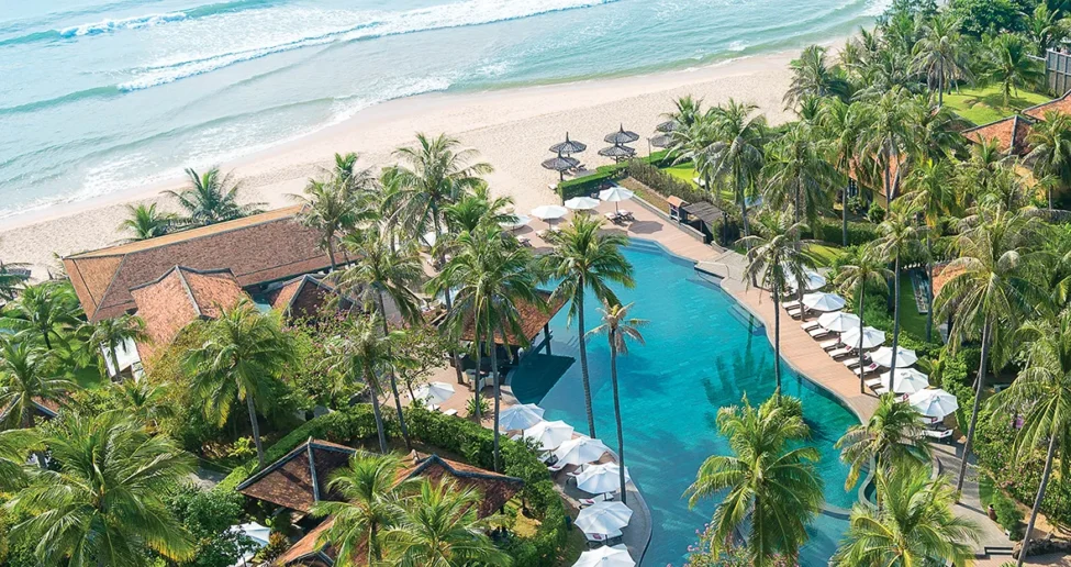 Luftaufnahme eines tropischen Strandresorts mit einem großen Pool, umgeben von Palmen, Liegestühlen, Sonnenschirmen und Gebäuden im traditionellen Stil, direkt an einem Sandstrand und den Wellen des Meeres.