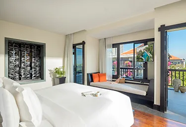 Anantara Mui Ne Deluxe Garten Zimmer Ein helles Hotelzimmer verfügt über ein großes Bett mit weißer Bettwäsche, eine Fensterbank mit orangefarbenen Kissen und Glasschiebetüren, die auf einen Balkon mit Topfpflanzen und Blick auf die bunten Dächer führen.
