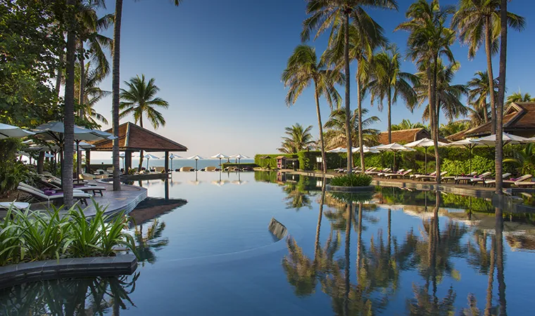 Anantara Mui Ne Pool Ein tropischer Resort-Pool, umgeben von Palmen und Liegestühlen, mit strahlend blauem Himmel und dem Meer im Hintergrund.