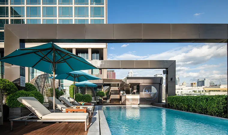Ein Dachpool mit Liegestühlen und türkisfarbenen Sonnenschirmen, umgeben von Grünflächen und modernen Metallkonstruktionen, unter strahlend blauem Himmel mit den Gebäuden der Stadt im Hintergrund.