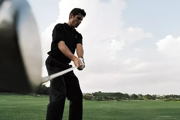 golfreise-mit-platzreife Ein Mann in schwarzer Kleidung macht sich bereit, auf einem grünen Golfplatz mit einem Driver einen Golfball zu schlagen; im Hintergrund sind Bäume und ein bewölkter Himmel zu sehen. Das Bild wurde aus einem niedrigen Winkel in der Nähe des Schlägers aufgenommen.