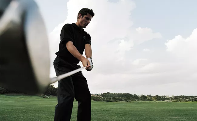 Ein Mann in schwarzer Kleidung macht sich bereit, auf einem grünen Golfplatz mit einem Driver einen Golfball zu schlagen; im Hintergrund sind Bäume und ein bewölkter Himmel zu sehen. Das Bild wurde aus einem niedrigen Winkel in der Nähe des Schlägers aufgenommen.