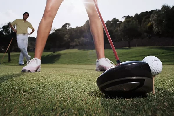 golfschlaege Nahaufnahme eines Golfschlägers und eines Balls auf einem Tee, im Vordergrund die Beine eines Golfers und im Hintergrund eine weitere Person, die unter strahlendem Himmel auf dem Golfplatz steht.