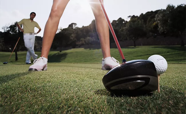 Nahaufnahme eines Golfschlägers und eines Balls auf einem Tee, im Vordergrund die Beine eines Golfers und im Hintergrund eine weitere Person, die unter strahlendem Himmel auf dem Golfplatz steht.