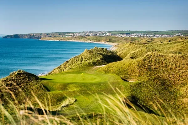 links-course-ballybunion Ein malerischer Links-Golfplatz besticht durch grüne Fairways und sanfte Sanddünen mit Blick auf das blaue Meer; in der Ferne ist unter dem klaren Himmel eine Stadt an der Küste zu sehen.