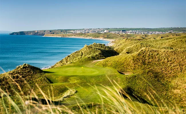 Ein malerischer Links-Golfplatz besticht durch grüne Fairways und sanfte Sanddünen mit Blick auf das blaue Meer; in der Ferne ist unter dem klaren Himmel eine Stadt an der Küste zu sehen.