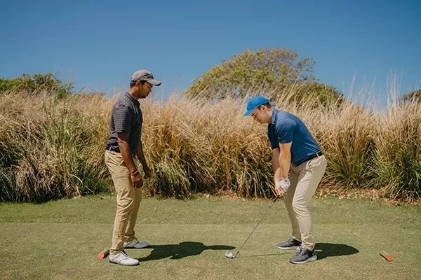 reisebericht-mauritius Zwei Männer stehen auf einem Golfplatz; einer hält einen Golfschläger in der Hand und macht sich bereit zum Schlag, während der andere zusieht. Im Hintergrund sind hohes Gras und Bäume zu sehen, über ihnen ein strahlend blauer Himmel.