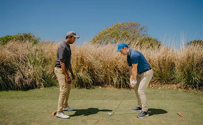 Zwei Männer stehen auf einem Golfplatz; einer hält einen Golfschläger in der Hand und macht sich bereit zum Schlag, während der andere zusieht. Im Hintergrund sind hohes Gras und Bäume zu sehen, über ihnen ein strahlend blauer Himmel.