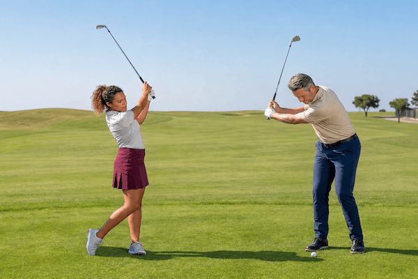 pitch-golf-schwung Zwei Personen üben auf einem Golfplatz ihren Schwung. Die Frau auf der linken Seite trägt ein weißes Shirt und einen roten Rock, während der Mann auf der rechten Seite ein beiges Shirt und eine blaue Hose trägt. Beide halten Golfschläger in der Hand, hinter ihnen erstreckt sich eine Wiese.
