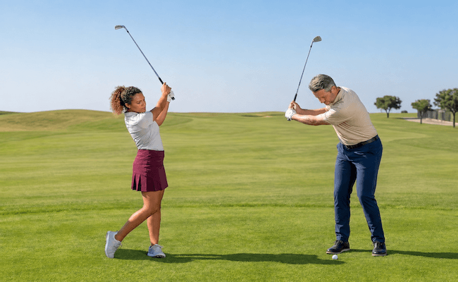 Zwei Personen üben auf einem Golfplatz ihren Schwung. Die Frau auf der linken Seite trägt ein weißes Shirt und einen roten Rock, während der Mann auf der rechten Seite ein beiges Shirt und eine blaue Hose trägt. Beide halten Golfschläger in der Hand, hinter ihnen erstreckt sich eine Wiese.