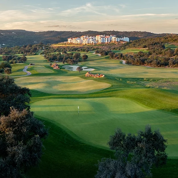 Weitwinkelansicht eines üppig grünen Golfplatzes mit Sandbunker, gewundenen Wegen und Bäumen. Im Hintergrund sind Hügel und eine Gruppe von Gebäuden – darunter das „Viceroy at Ombria Algarve“ – zu sehen, vor einem teilweise bewölkten Himmel bei Sonnenuntergang.