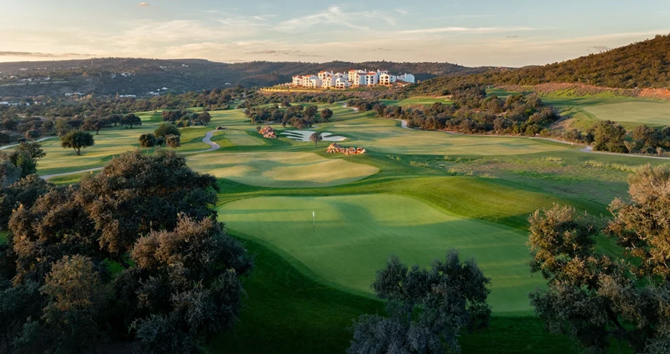 Weitwinkelansicht eines üppig grünen Golfplatzes mit Sandbunker, gewundenen Wegen und Bäumen. Im Hintergrund sind Hügel und eine Gruppe von Gebäuden – darunter das „Viceroy at Ombria Algarve“ – zu sehen, vor einem teilweise bewölkten Himmel bei Sonnenuntergang.