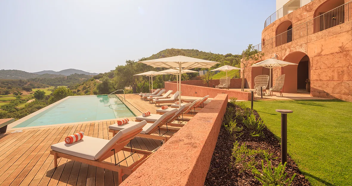 Viceroy at Ombria Algarve_Pool Der luxuriöse Poolbereich mit Liegestühlen, Sonnenschirmen und gestreiften Handtüchern bietet im „Viceroy at Ombria Algarve“ einen Blick auf grüne Hügel und einen Golfplatz, direkt neben einem terrakottafarbenen Gebäude unter strahlend blauem Himmel.