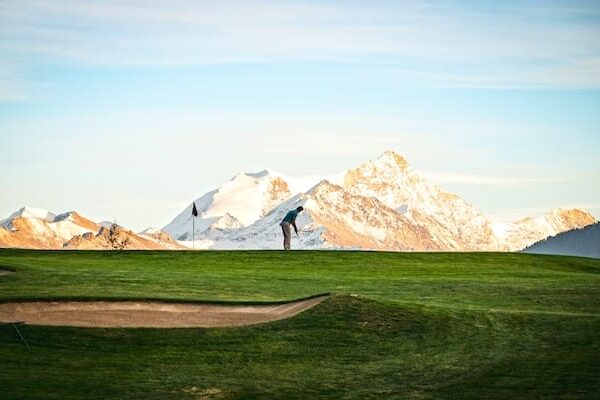 annaeherungsschlag-golf Ein Golfer puttet auf einem Grün, im Hintergrund sind schneebedeckte Berge zu sehen, über ihnen ein teilweise bewölkter Himmel.