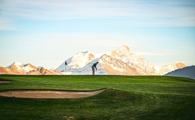 annaeherungsschlag-golf Ein Golfer puttet auf einem Grün, im Hintergrund sind schneebedeckte Berge zu sehen, über ihnen ein teilweise bewölkter Himmel.