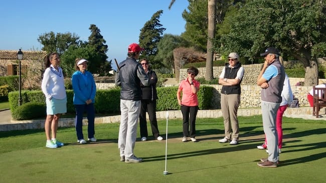annaeherungsschlag-golflehrer Eine Gruppe von Menschen, von denen einige Golfschläger in der Hand halten, steht im Kreis auf einem Putting Green und hört einer Person mit roter Mütze zu. Im Hintergrund sind Bäume und Steingebäude zu sehen.