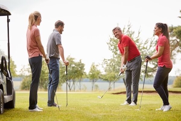 Golf Professional Demonstrating Shot On Fairway To Group Of Golfers During Lesson Vier Personen stehen mit Golfschlägern auf einem Golfplatz, lächeln und unterhalten sich. In der Nähe steht ein Golfwagen, und im Hintergrund sind Bäume zu sehen.