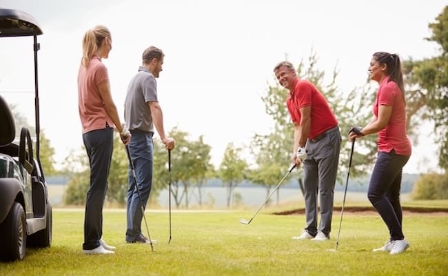 Vier Personen stehen mit Golfschlägern auf einem Golfplatz, lächeln und unterhalten sich. In der Nähe steht ein Golfwagen, und im Hintergrund sind Bäume zu sehen.