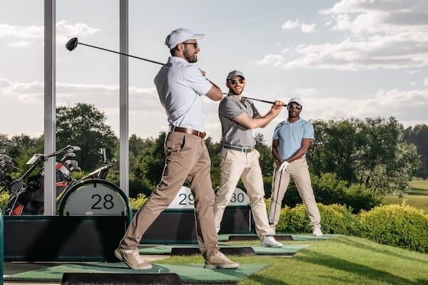 group of golf players playing golf together at golf course Drei Männer in Golfkleidung stehen auf einer Driving Range. Zwei schwingen ihre Schläger, während der dritte seinen Schläger in der Hand hält und zusieht. Im Hintergrund sind unter einem teilweise bewölkten Himmel Golftaschen und grüne Bäume zu sehen.
