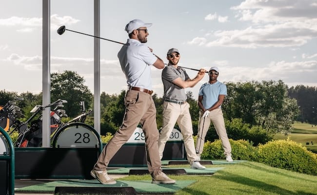 group of golf players playing golf together at golf course Drei Männer in Golfkleidung stehen auf einer Driving Range. Zwei schwingen ihre Schläger, während der dritte seinen Schläger in der Hand hält und zusieht. Im Hintergrund sind unter einem teilweise bewölkten Himmel Golftaschen und grüne Bäume zu sehen.