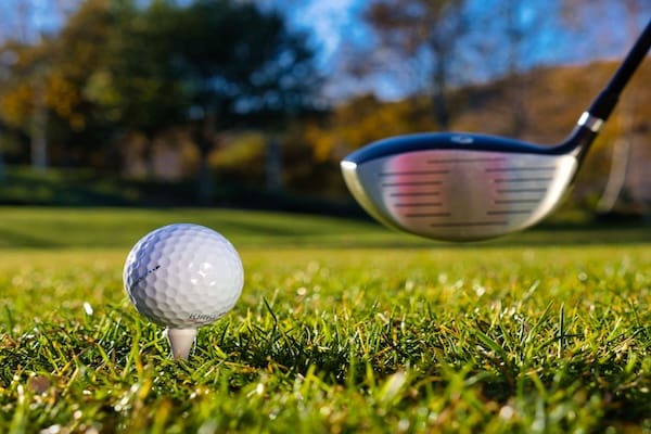 golf-abschlag Nahaufnahme eines Golfballs auf einem Tee im Gras, während ein Golfschläger kurz davor ist, ihn zu treffen. Im Hintergrund sind verschwommene Bäume und ein blauer Himmel an einem sonnigen Tag zu sehen.
