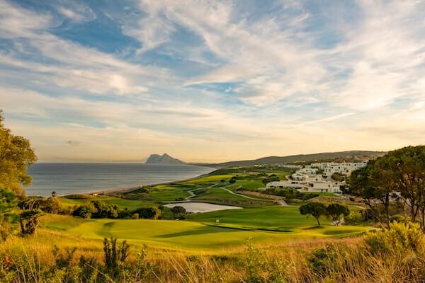 Ein Golfplatz an der Küste mit grünen Fairways, weißen Gebäuden und dem Meer auf der linken Seite. Unter einem blauen Himmel mit vereinzelten Wolken sind in der Ferne Berge zu sehen.