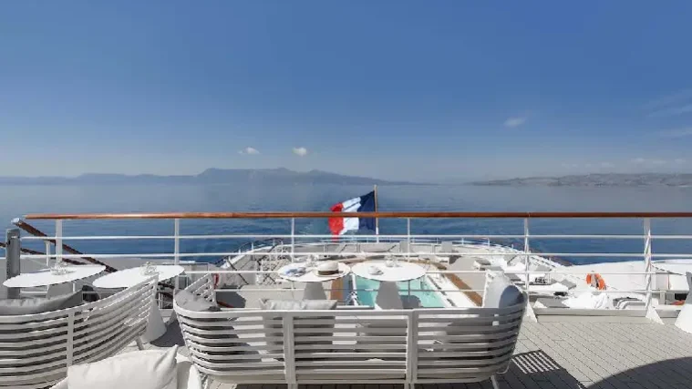 Der Blick vom Deck einer Yacht mit weißen Sitzgelegenheiten auf den Pool und das Meer vermittelt den luxuriösen Charme des „Golf-Frühlings“ in Costa Navarino. Am Heck weht eine französische Flagge vor dem Hintergrund der Berge und des strahlend blauen Himmels.