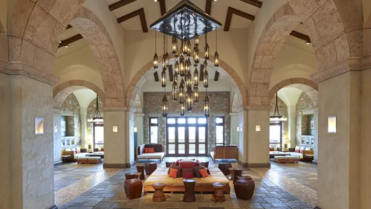 Eine großzügige Hotellobby mit Steinbögen, Hängeleuchten und großen Fenstern heißt die Gäste zum Saisonauftakt in Costa Navarino willkommen. In der Mitte, auf einem mit Steinfliesen ausgelegten Boden, sind moderne Holzsitzgelegenheiten mit roten Kissen angeordnet, während vom Eingang aus Licht hereinströmt.